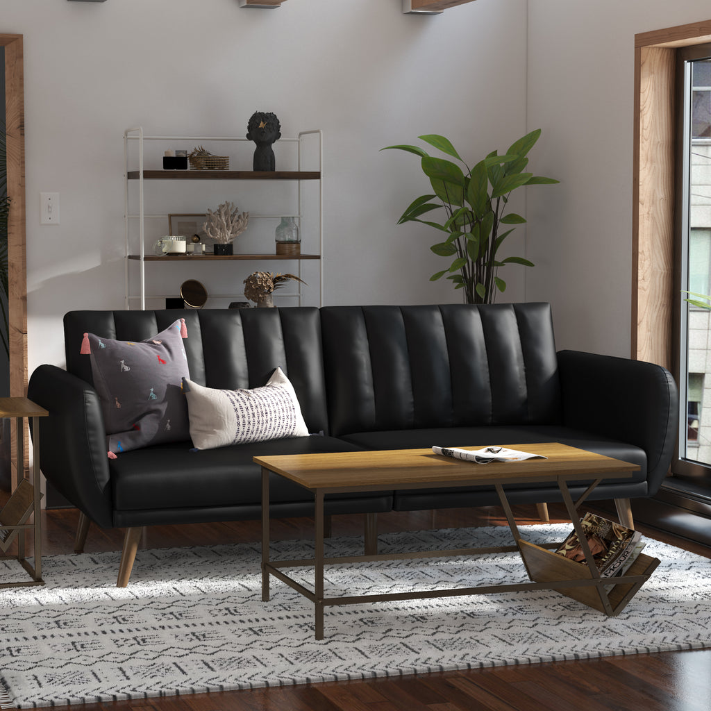 A modern living room showcases the Brittany Futon in sleek black faux leather with patterned cushions, featuring a split-back design. A wooden coffee table is placed on a geometric rug, while a metal shelf decorated with decor and a potted plant complements the sunlit space.