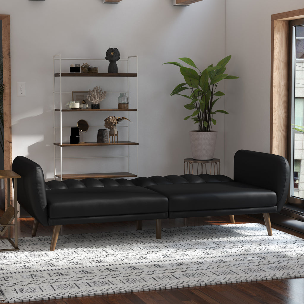 A modern living room features a Brittany Futon - Faux Leather with wooden legs on a patterned rug. Sunlight illuminates the potted plant beside it, and a shelving unit in the corner displays decorative items.