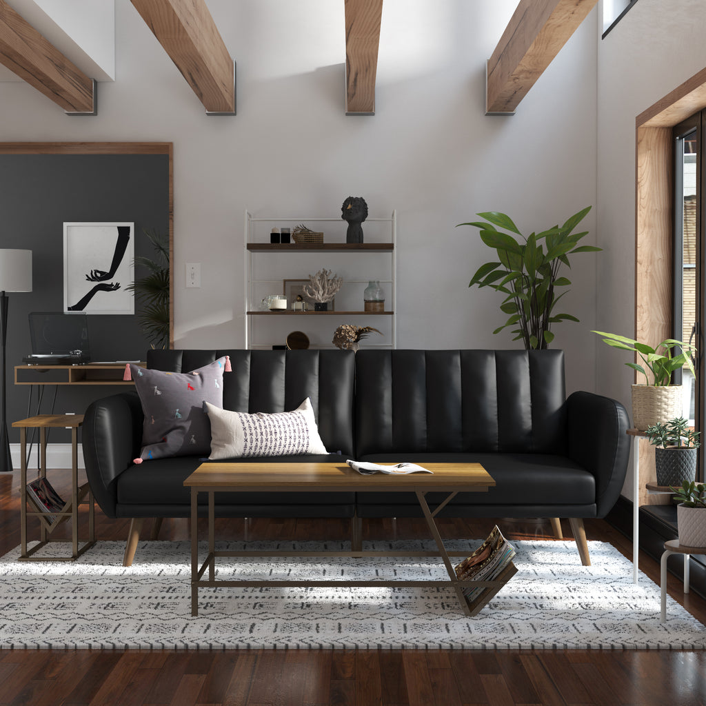 A modern living room showcases the Brittany Futon in sleek black faux leather, adorned with patterned cushions. A wooden coffee table with metal legs complements it, sunlight pours through a window, and décor includes a bookcase, plants, and minimalist art, exuding contemporary charm.