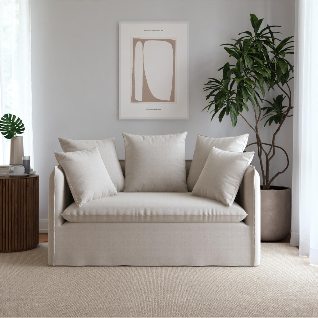 A minimalist living room showcases a Collette Slipcover Loveseat Sofa, in beige, with matching cushions. To the right sits a large potted plant, while a wooden side table with a small plant is on the left. The wall behind is elegantly adorned with simple abstract artwork.