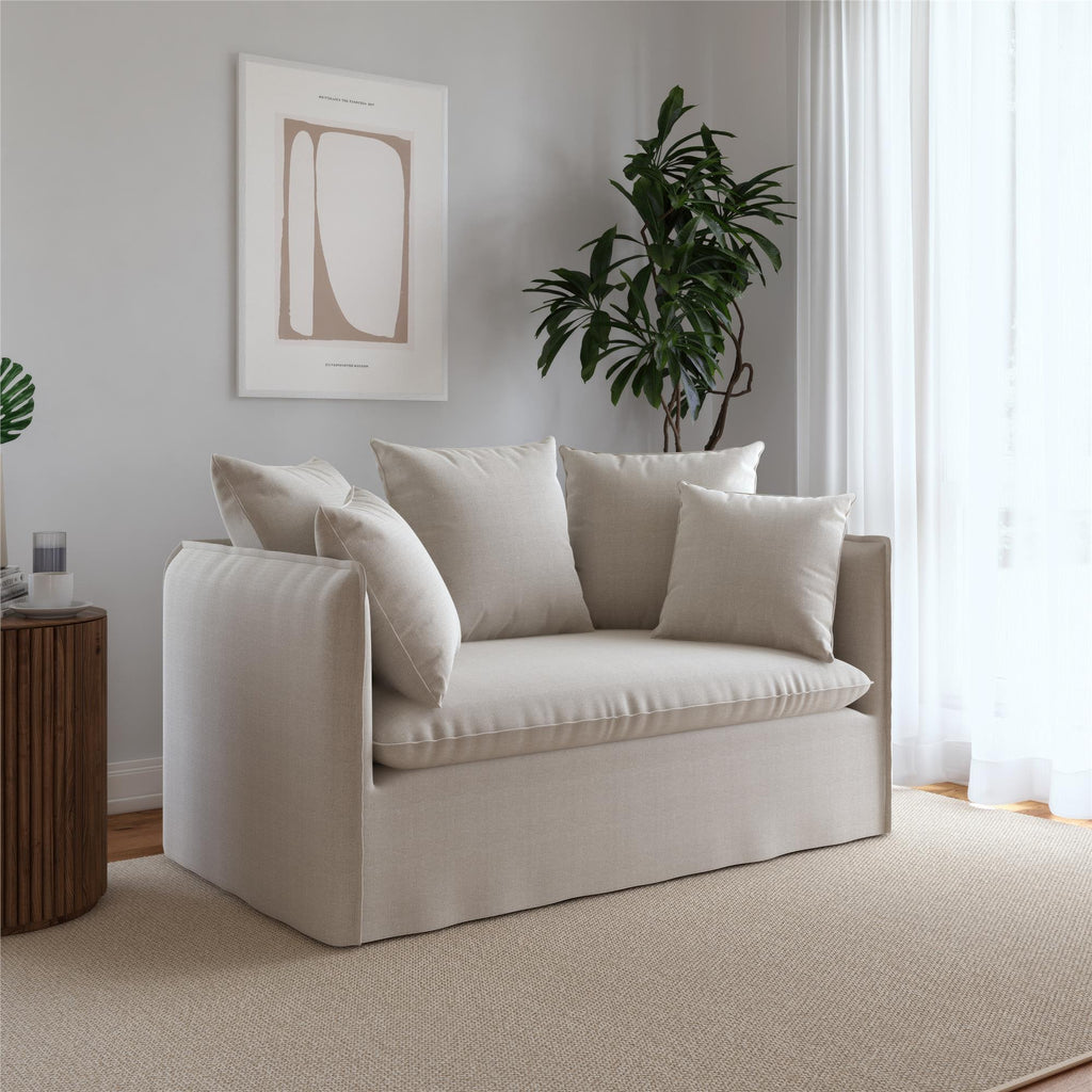 The Collette Slipcover Loveseat Sofa in light beige, featuring removable slipcovers and multiple cushions, sits in a modern living room. A potted plant is nearby, while minimalist abstract art adorns the wall, and natural light streams through sheer curtains.