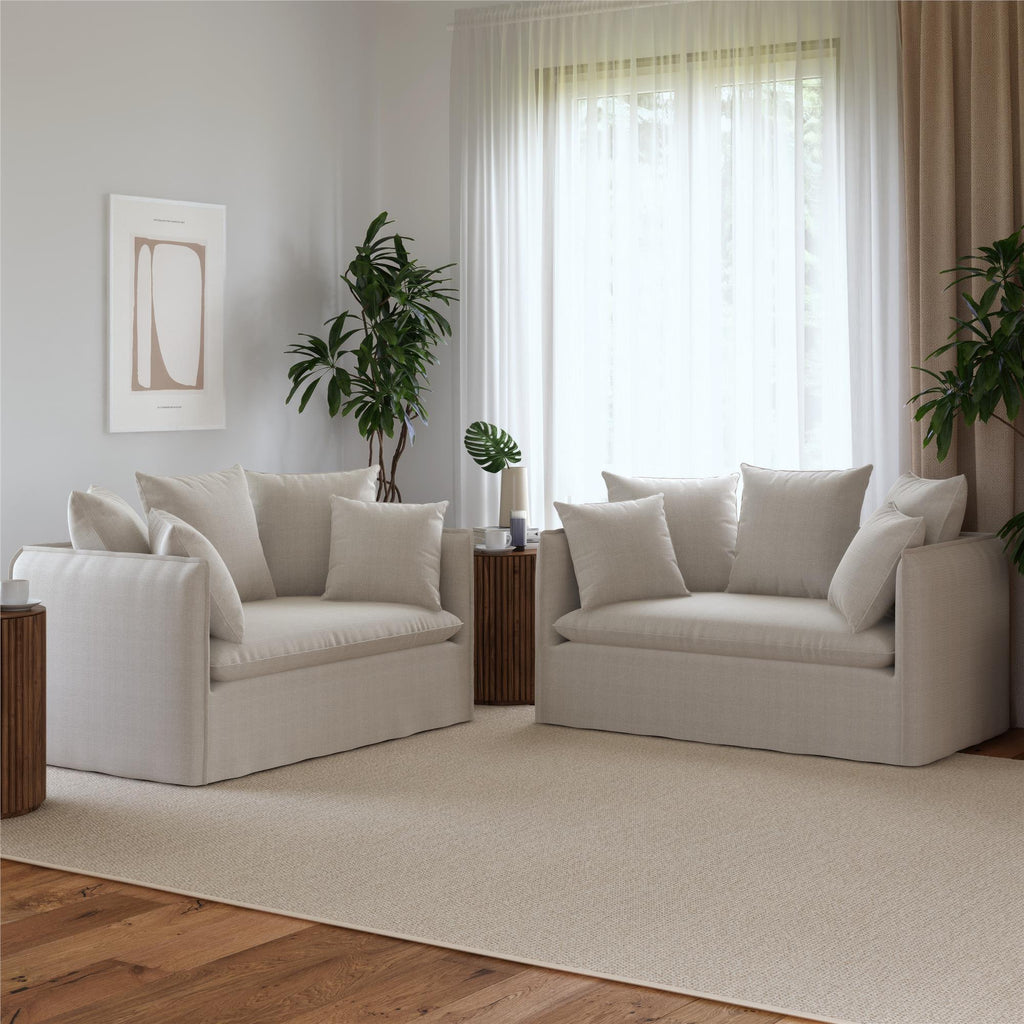 A minimalist living room features a Collette Slipcover Loveseat Sofa and a matching one, both adorned with pillows. Light floods in through sheer curtains on a large window. High-density foam seating ensures comfort, while two potted plants sit between tables, and abstract art enhances the wall.