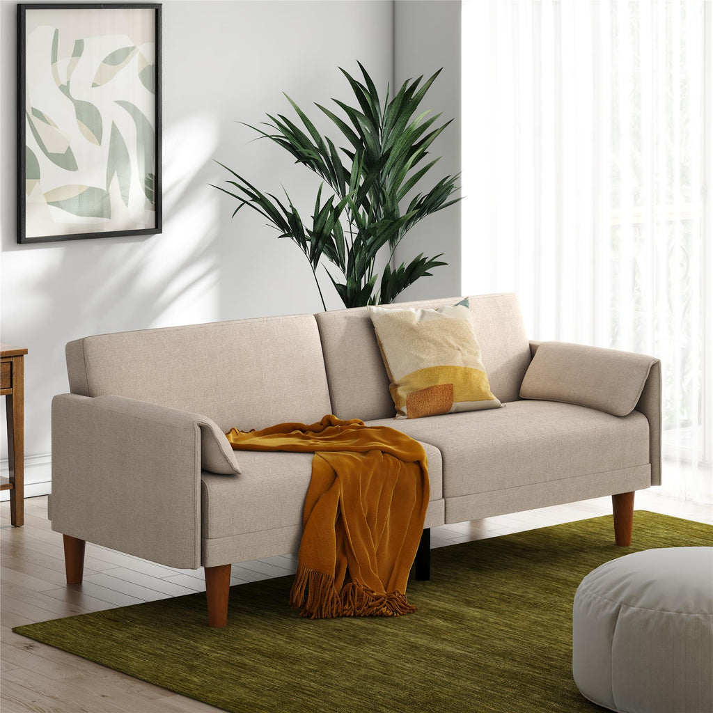 The beige Markham Futon with wooden legs is decorated with a yellow pillow and an orange throw blanket. A tall green plant stands nearby, complemented by minimalist leaf artwork on the wall. Sheer curtains allow light to brighten a green rug.