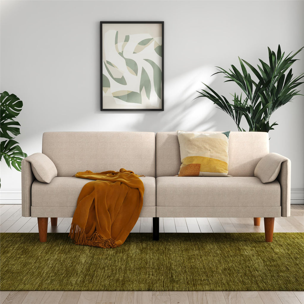 A minimalist living room showcases the Markham Futon with a yellow blanket and multi-colored pillow. A framed leafy art piece hangs above, with two potted plants flanking the futon and a green rug adorning the light wood floor.