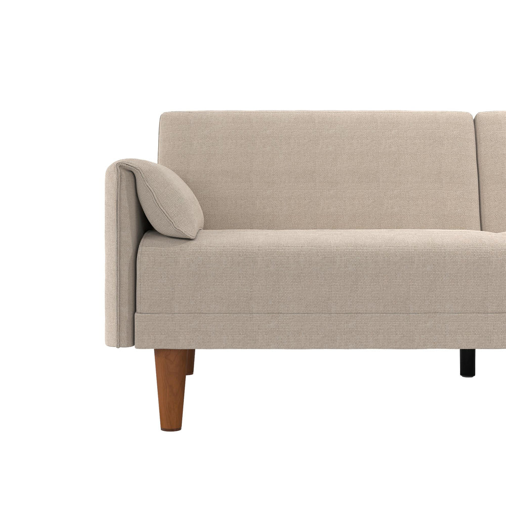 The Markham Futon is a light beige Scandinavian sleeper sofa boasting a minimalist design, a cushioned back, and a left-side armrest. It stands on visible wooden legs, one light and one dark, set against a plain white background.