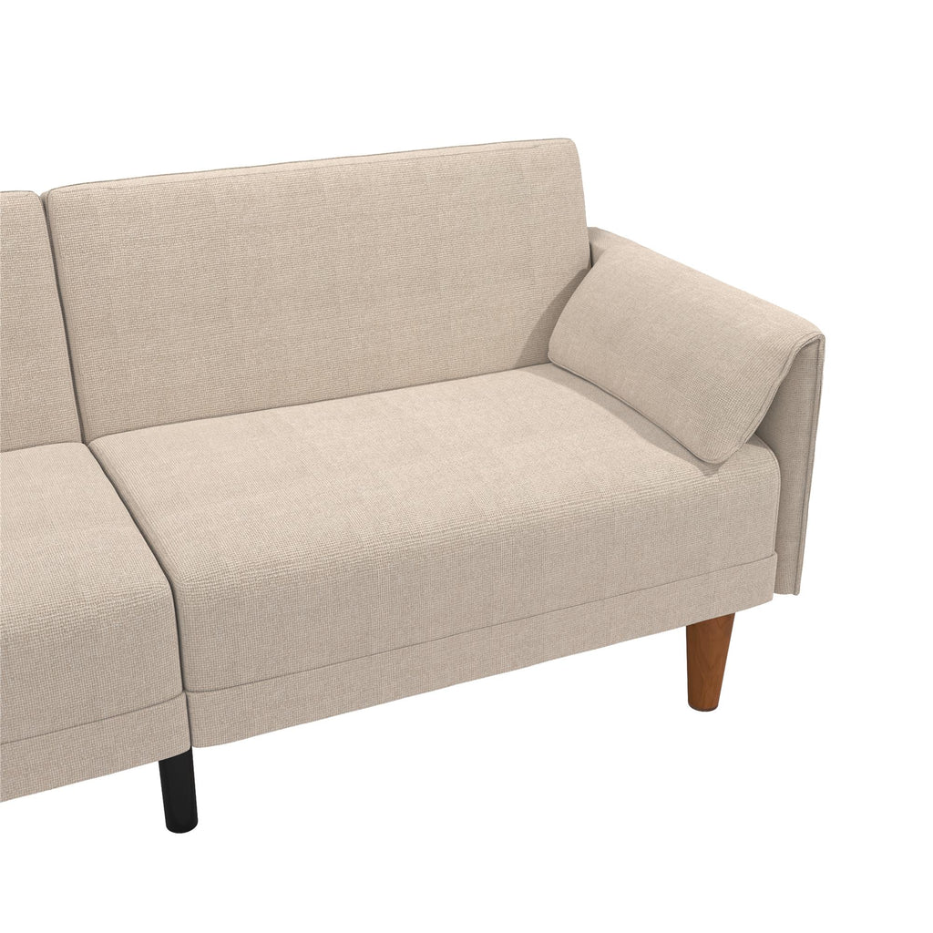 The Markham Futon is a beige sectional sofa with minimalist Scandinavian design, showcasing clean lines and wooden legs. The right side has an armrest and reclined backrest, while the left part is partially visible against a plain white background.