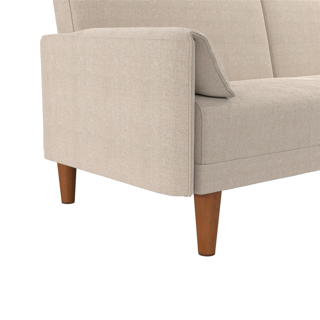 A close-up of the Markham Futon, a beige Scandinavian sleeper sofa with wooden legs, highlights its clean, modern design. The textured fabric emphasizes intricate upholstery details and showcases soft armrests and back cushions.