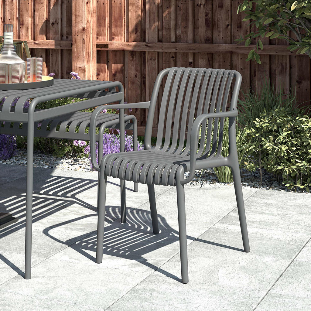 A set of Bennett Stacking Dining Chairs with a gray metal, slatted design sits on a stone patio beside the matching table. A wooden fence and plants are visible in the background on this sunny day. Enjoy effortless style with no assembly required.