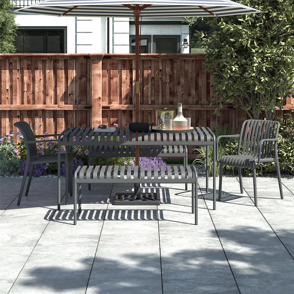 A patio with a striped umbrella shades a table, two Bennett Stacking Dining Chairs from a set of four, and a bench. The table holds a bottle and glasses. The area is paved with large tiles and surrounded by a wooden fence and lush greenery.