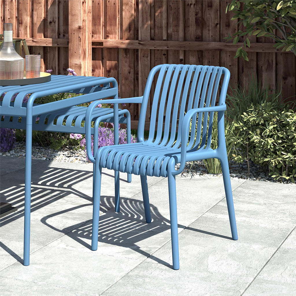 A blue metal chair, echoing the Bennett Stacking Dining Chairs (Set of 4) with its slatted design, sits on a stone patio beside a matching table. A tray with a carafe and glasses completes the setting framed by a wooden fence, plants, and vibrant flower beds.