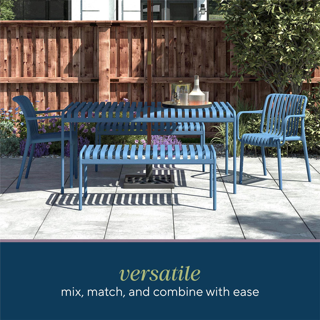 A patio set on tiles includes durable Bennett Stacking Dining Chairs (Set of 4), a bench, table, and a parasol at the center. A wooden fence and greenery form a calming backdrop. Text: Versatile - easily mix, match, combine; no assembly needed.