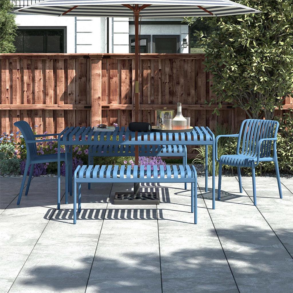 The outdoor patio scene features a blue-striped metal table and Bennett Stacking Dining Chairs (Set of 4) under a striped umbrella. A wooden fence and greenery frame the background, while sunlight highlights a water bottle, tray, and small speaker. The chairs are durable, stylish, and come fully assembled.