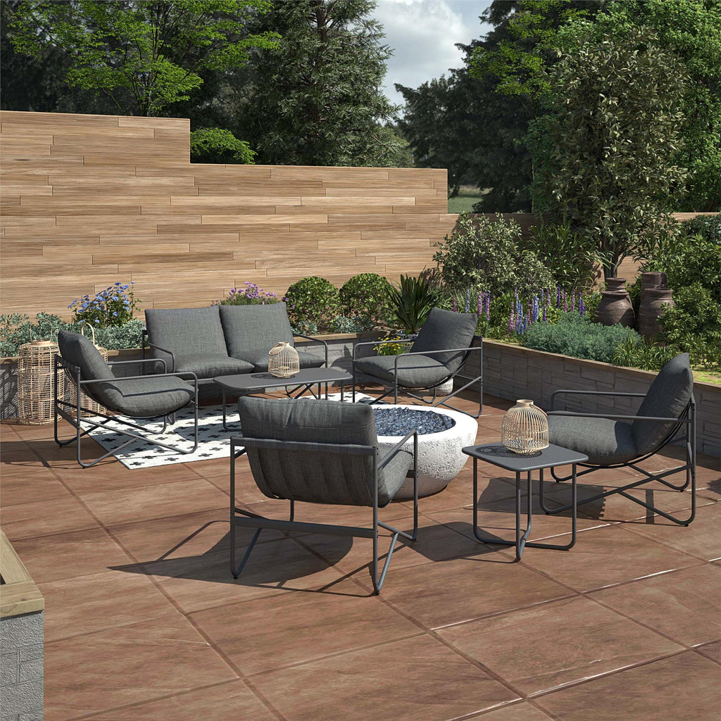 The tiled outdoor patio features the Wrenn 4-Piece Sling and Cushion Conversation Set with gray cushioned seating around a round concrete fire pit. A small table displays a decorative item, while planters with lush greenery, wooden panel walls, and trees create the perfect setting for outdoor gatherings.