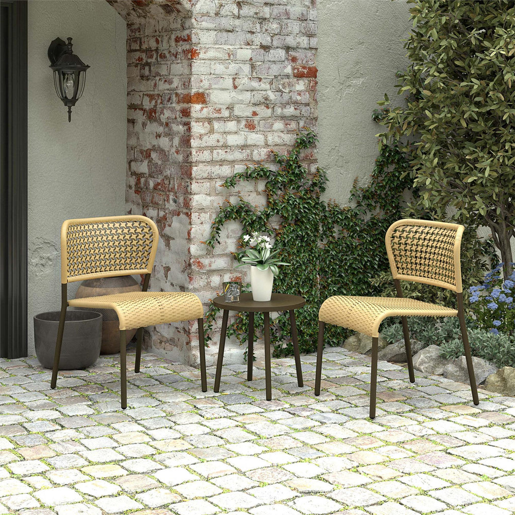 The Briar 3-Piece Wicker Chat Set, with two chairs and a round table adorned by a potted plant, sits on a cobblestone patio. Against a rustic brick wall with climbing greenery, it offers cozy outdoor seating that blends modern design with nature.