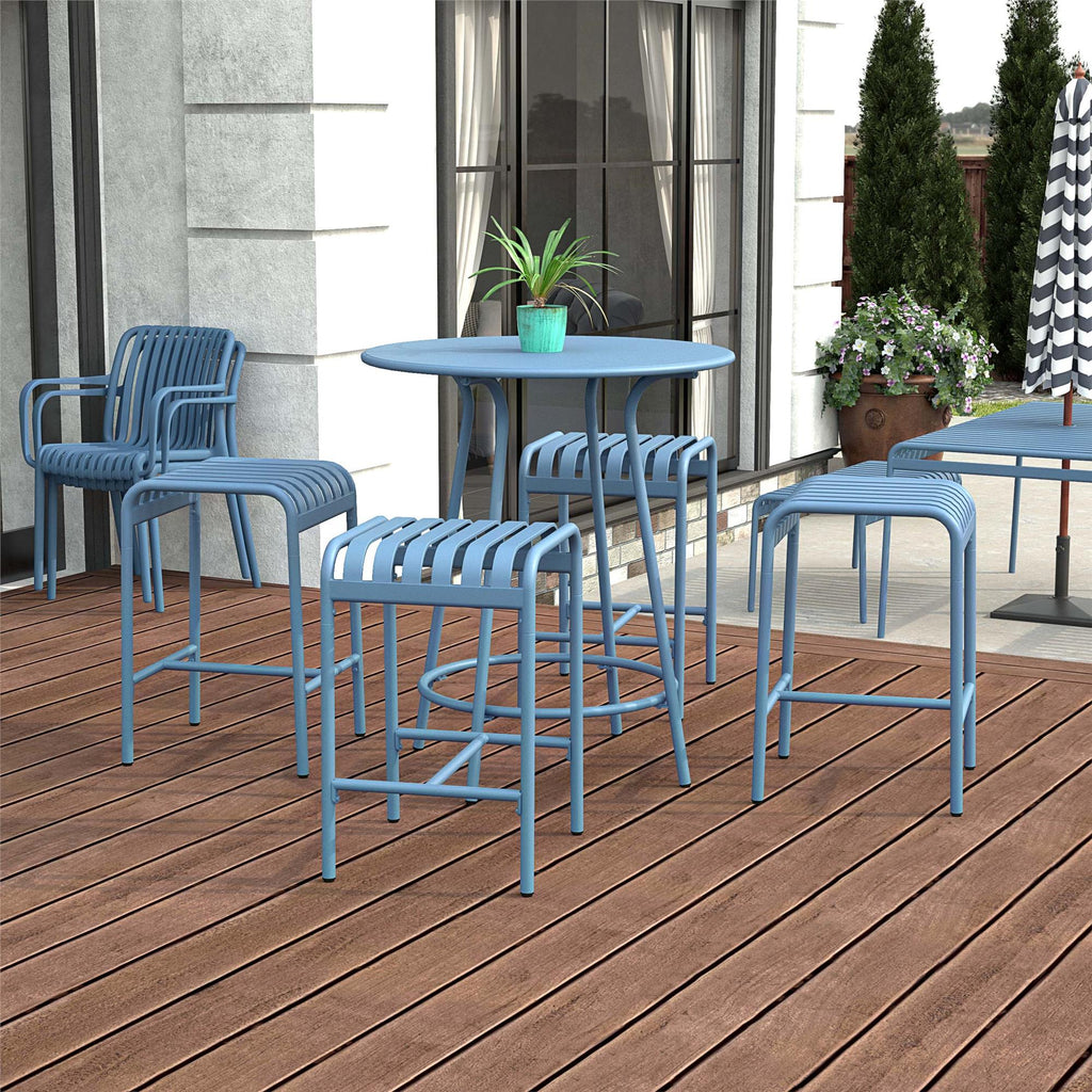 The Ola 5-Piece High Dining Set features a blue outdoor patio setup with durable steel components, providing cozy seating for four chairs and two stools. A small potted plant adorns the table, complemented by a large window backdrop, building elements, and lush greenery.