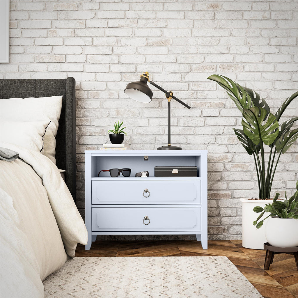 A cozy bedroom features a white brick wall and a large bed with white linens. The Her Majesty 2 Drawer Nightstand in light blue elegantly holds a lamp, eyeglasses, and a small plant, complementing the tall green plant on the parquet floor.