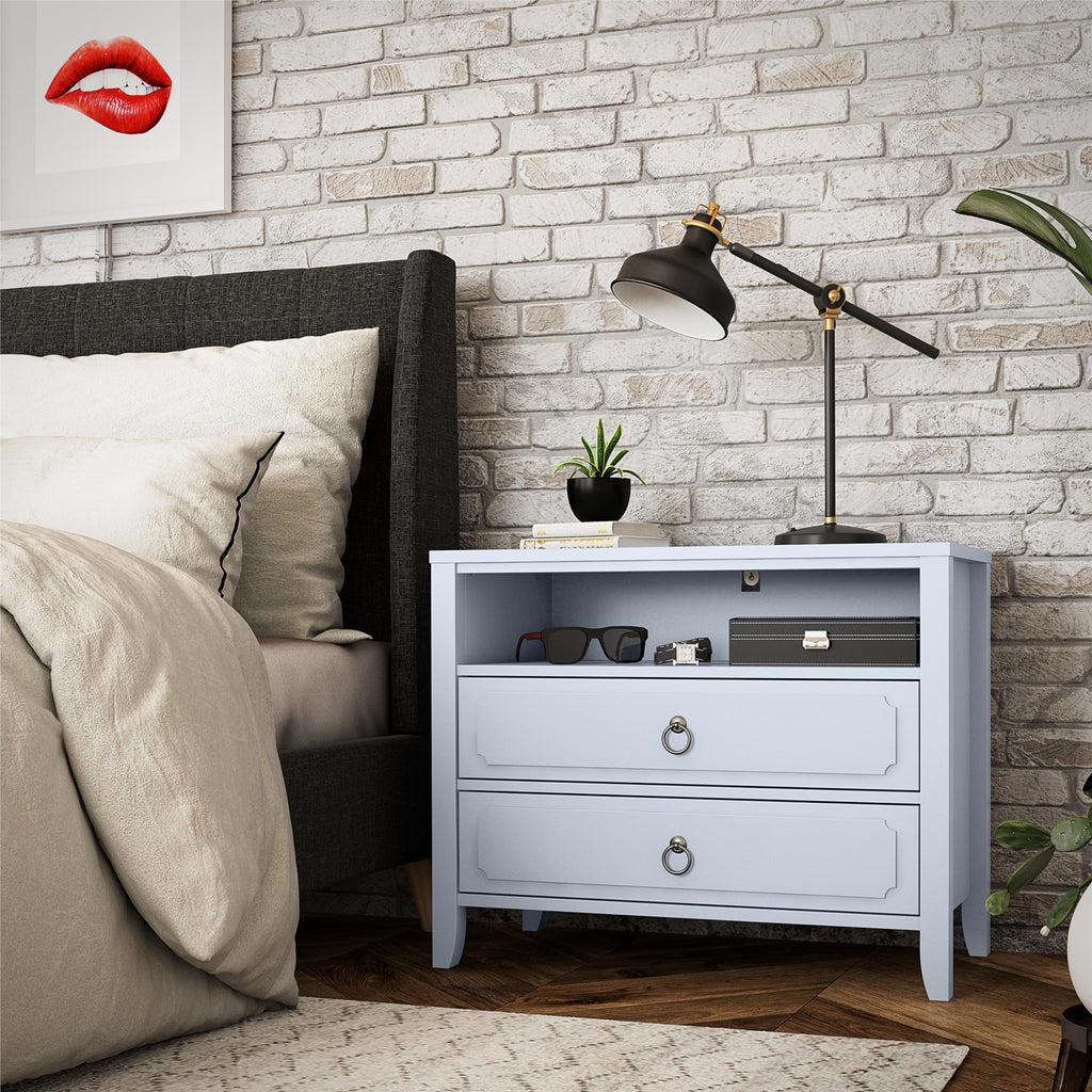 A cozy bedroom scene features a grey bed with beige bedding beside the elegant light blue Her Majesty 2 Drawer Nightstand from Novogratz, holding a lamp, small plant, and glasses. An eye-catching red lips artwork adorns the brick wall above.