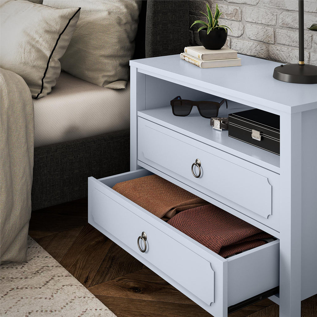 The Her Majesty 2 Drawer Nightstand, in elegant light blue, stands beside the bed with two drawers. The top drawer is slightly open, revealing brown fabric inside. Its surface holds a small plant, books, sunglasses, and a black box that complements the beige and white bedding.