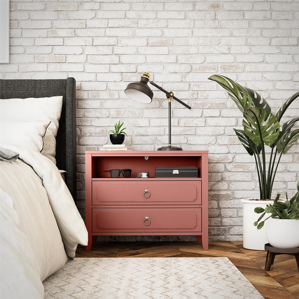 A bedroom scene features an elegant Her Majesty 2 Drawer Nightstand adorned with a black and gold lamp, potted plants, and small items. The red nightstand stands next to a bed with white bedding against a white brick wall on a wooden floor.