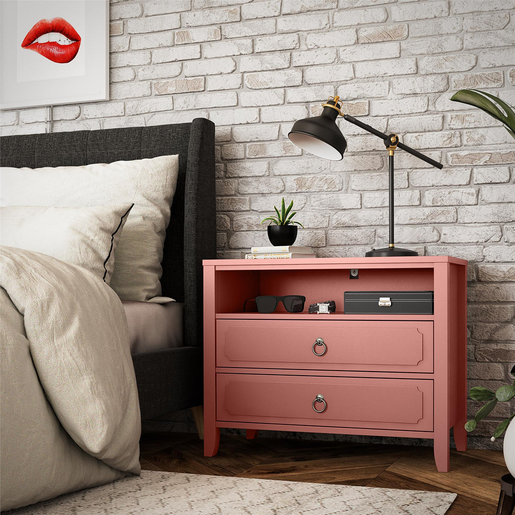 A cozy bedroom corner features a pink Her Majesty 2 Drawer Nightstand by Novogratz, complete with a black lamp, glasses, plant, and small box. The gray bed with white bedding is set against a brick wall adorned with a framed picture of red lips above.