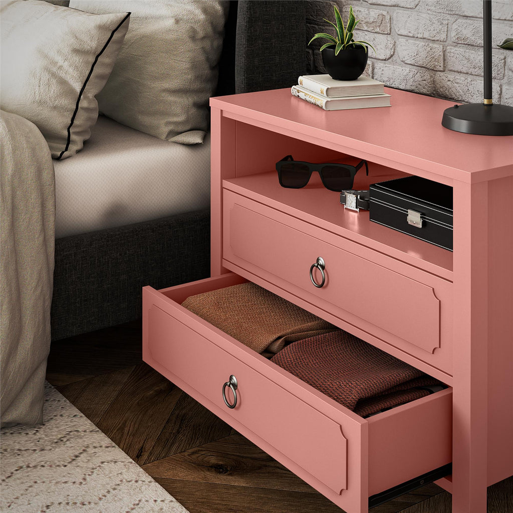 The Her Majesty 2 Drawer Nightstand, in elegant pink, boasts two open drawers. The top shelf holds sunglasses, a black box, books, and a potted plant. Nearby is a cozy bed with gray bedding on a textured rug against a white brick wall.