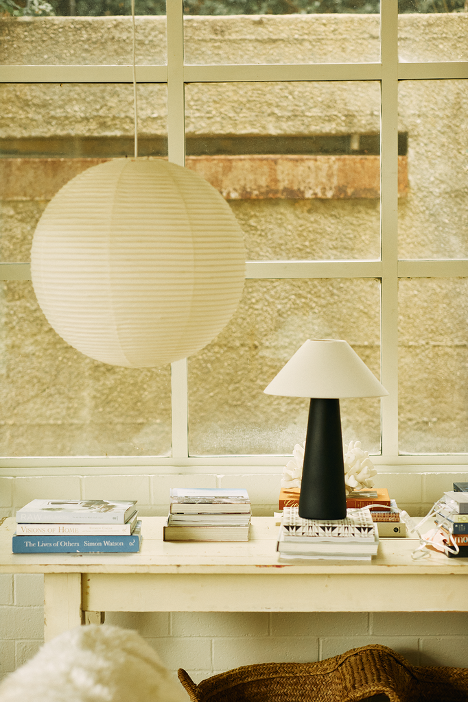 A cozy room features a large paper lantern on the ceiling and a Hutton 18 Table Lamp with a black base and white shade lighting up a book-stacked table. A large window offers an outside view, beautifully mixing warmth with industrial design elements.