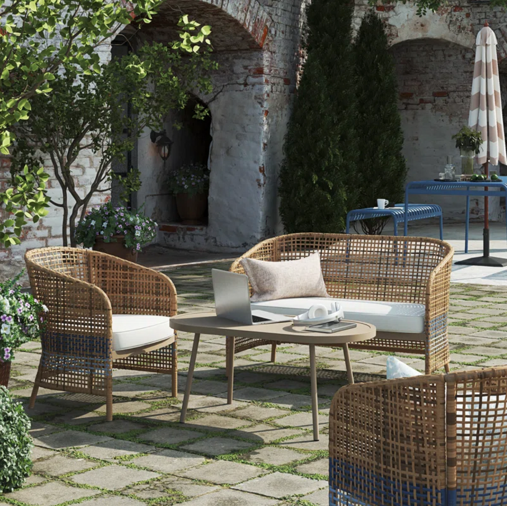 The outdoor patio features the Ava 4-Piece Wicker Conversation Set in French blue, with plush chairs and a loveseat surrounding a wooden coffee table. A laptop and headphones are on the table. Stone paving complements lush greenery, with a white brick wall as an elegant backdrop.