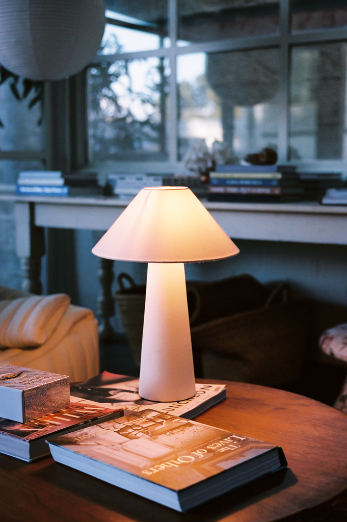 A cozy table setting features a Hutton 18 Table Lamp casting a warm glow, complemented by stacked books on wood. An inviting sofa with striped pillows and a side table filled with more books completes the peaceful reading nook ambiance.